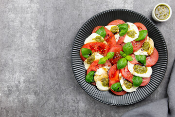 Traditional italian caprese salad with sliced tomatoes, mozzarella, basil, pesto sauce and spice on a dark background. Top view. Copy space.