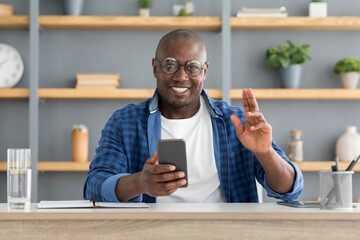 Cheerful mature african american man using smartphone and making greeting gesture, looking and smiling to camera