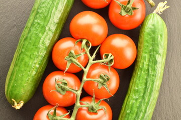 Several green cucumbers and red ripe, cocktail tomatoes on a twig, close-up, on a slate board, top view.