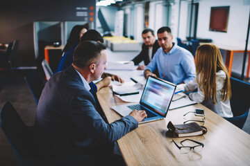 Formally dressed businessman making online booking and banking via laptop application while blurred colleagues cooperating, executive managers searching financial website for checking trade exchange