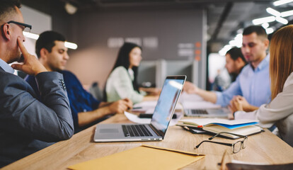 Group of productive employees collaborating at desktop in office discussing ideas for startup, male and female partners talking about trade and project analyzing details during briefing meeting