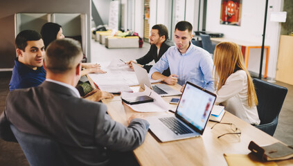 Group of productive employees collaborating at desktop in office discussing ideas for startup, male and female partners talking about trade and project analyzing details during briefing meeting