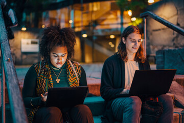 Young male and female students using laptop while sitting on steps in campus at night