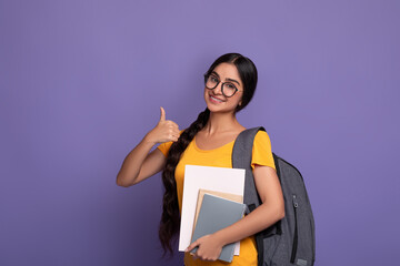 Happy indian student wearing eyeglasses holding notebooks showing thumbs up