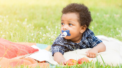 Cute little dark skinned, mix race African baby boy with pacifier in the mouth, lying on the rug at picnic in the park.