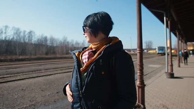 Woman wearing glasses sling jacket waiting for the train at the station. Rotation of the camera in a circle. Sunlight.