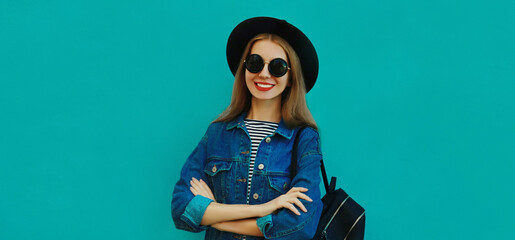 Portrait close up of smiling young woman wearing a black round hat, denim jacket and backpack on a blue background