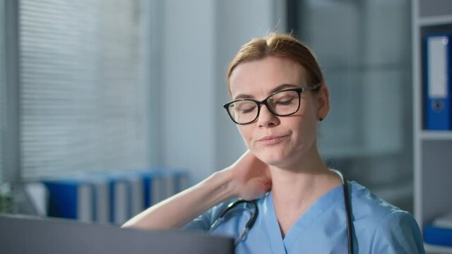 doctor while working feels pain in the neck, tired caucasian female healthcare professional in glasses wearing blue uniform looks at camera close-up