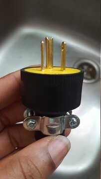 A hand holding a yellow and black three-prong electrical plug over a blurred silver kitchen- sinkhole. 