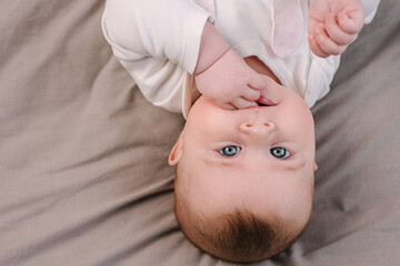 Top view portrait of cute charming baby girl holding hand in mouth lying on comfy bed at home look at camera posing. Little newborn infant child play in cozy bedroom. Upbringing, childcare concept.