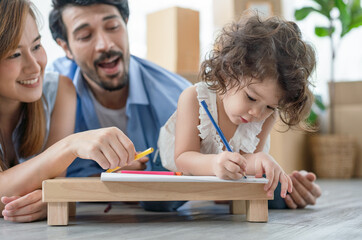 Happy family mother and father lying on the floor teaching their daughter drawing in color book together in living room at home