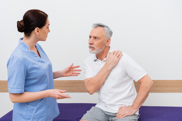 Obraz premium rehabilitologist gesturing while talking to mature man touching shoulder in clinic