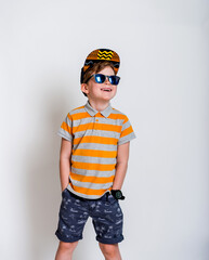 Fototapeta premium Cute stylish boy ready for summer. Seven years old boy with hands in pockets and yellow T-shirt is looking up. A merry cap and sunglasses. Standing on white background,