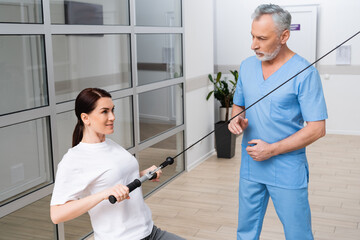 mature trainer assisting woman working out on pull rope exercising machine