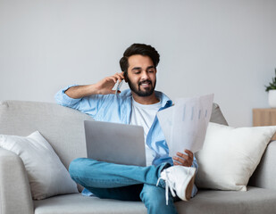 Eastern Freelancer Man With Cellphone, Papers And Laptop Working Remotely At Home