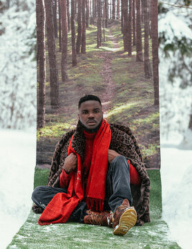A beautiful African-American man in a winter forest against the background of summer and green grass in stylish clothes. The concept of summer in winter.
