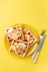 Waffles with banana, almond slices, cream and caramel sauce isolated on yellow background, top view. Sweet breakfast or dessert waffles