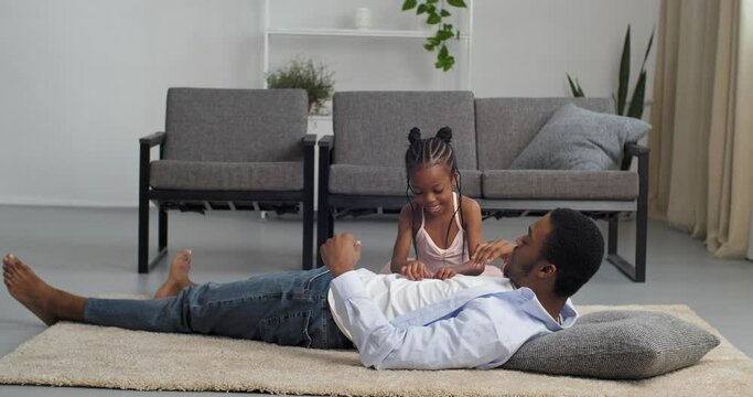 Afro american young father playing with black little girl daughter at home on floor, ethnic african man daddy lying in living room like patient in hospital child baby beats touching his belly abdomen