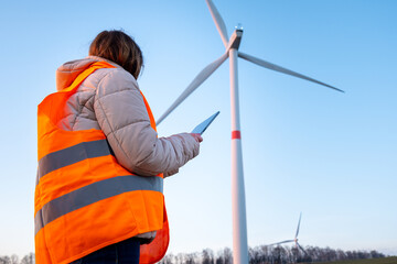 A woman engineer, holding a tablet and working at the wind turbine farm in the field in the orange...