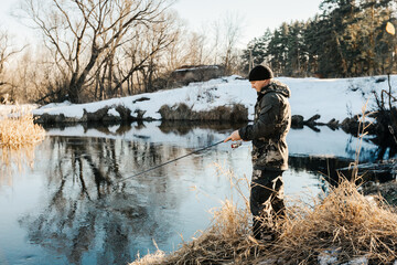A fisherman with a fishing rod catches fish on the bank of a snow-covered river in early spring