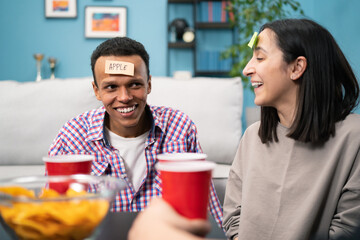 A group of friends are hanging out in the living room. A black american student boy plays who i am with his college friends with Sticky Papers Attached to Foreheads