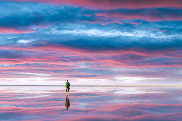 A tourist stands in calm water, which reflects the incredible purple sunset sky. Landscape...