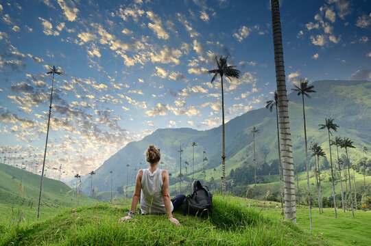  Girl with backpack sitting on green grass watching gigantic palm trees in Cocora Valley in Colombia