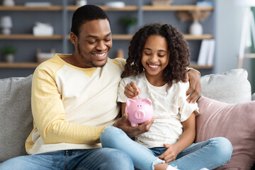 Black girl and father putting coin into piggy bank