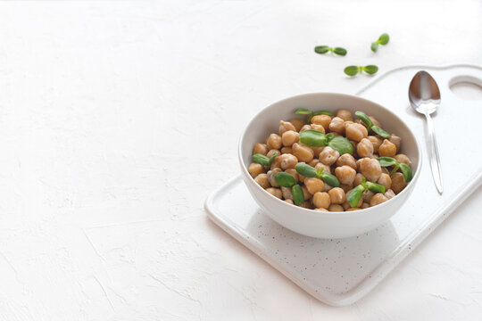 Cooked chickpeas with sunflower microgreen sprouts in a bowl on a cutting board on a white textured background