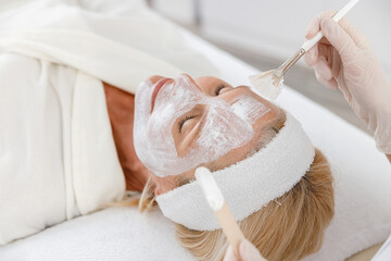 Close up photo of beautician applying nutrition face mask on senior woman face.