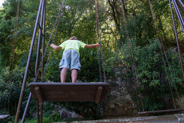 Naklejka na ściany i meble Boy is swinging on big swings in green summer park. Activities on vacation, happy childhood