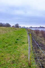 Fototapeta na wymiar Fence with barbed wire along the Maas river in a Dutch meadow with green grass and wil plants, cloudy day in Geulle in South Limburg, the Netherlands