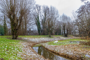 Stream with clean water and slight reflection among green grass with traces of snow, bare trees with some houses in the background in Kelmonderbos park on winter day in South Limburg, Netherlands