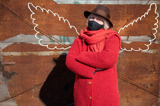 Grandma Is An Angel. Portrait Of A Fashionable Blond Old Lady In A Black Protective Mask Against A Rusty Wall With Painted Wings. Pandemic Covid-19 Coronavirus. Autumn Spring. Elderly Woman Fashion