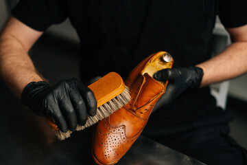 Close-up hands of shoemaker in black gloves polishing light brown leather shoes with brush during...
