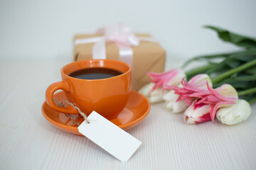 Romantic breakfast with orange coffee cup, gift box, pink tulips flowers on white background.Holiday mockup.