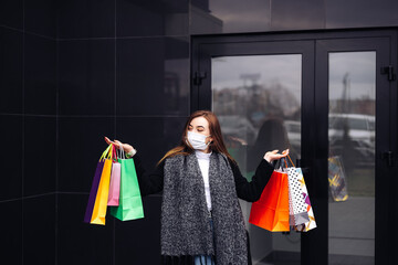 European lady hold hand colorful bags woman walking after shopping wear face blue disposable mask dressed warm coat scarf retail store near mall on the street sales black friday season upset


