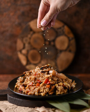 female hand sprinkles white sesame seeds on chahan rice with mussels and vegetables in a black bowl on a slate board with bamboo leaves on a brown background.