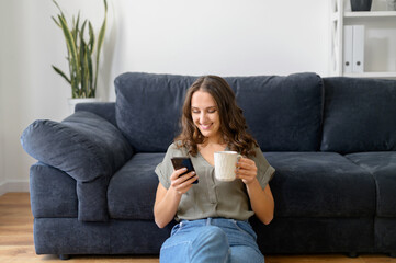 Pretty young woman using smartphone and drinking coffee sitting on the floor in cozy living room,...