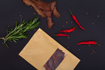 Jerky snacks, red papper, rosemary and craft package on black background. Mockup. Dried and spiced...