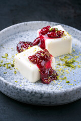 Modern style traditional blancmange almond pudding with sour cherry coulis and pistachios served as close-up in a Nordic design plate on black background