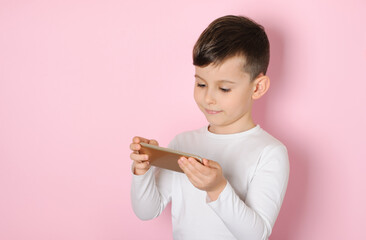 Little boy talking on the phone playing video games on a mobile phone wearing in a white t-shirt on a pink background. Distance learning during quarantine