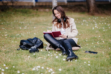 smiling female student learning remotely outdoors in park. Girl online learning courses using smartphone sitting in park. 