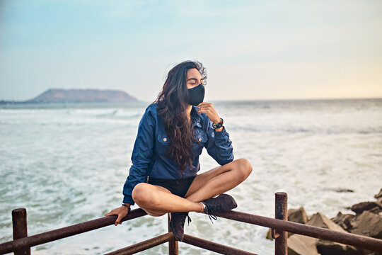 mujer joven con mascarilla feliz en la playa. Momento de relajacion a orillas del mar durante un atardecer. Fondo de olas y mar