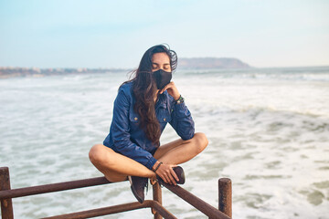 mujer joven con mascarilla feliz en la playa. Momento de relajacion a orillas del mar durante un atardecer. Fondo de olas y mar