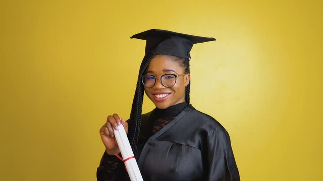 The african american graduate student in magister's robe stands on yellow background and holds in her hands a diploma of international standard of Ph.D. or Doctorate degrees