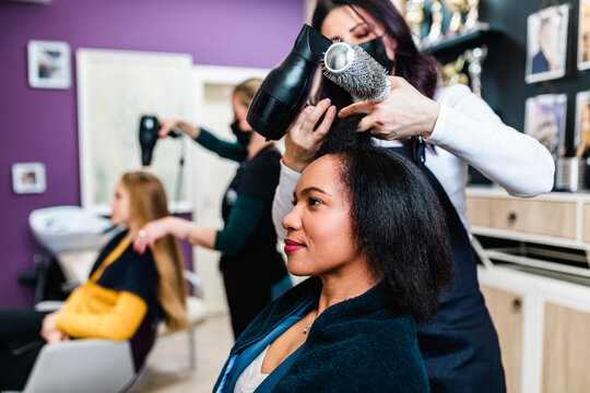 A hairdresser dries a Black woman's hair in a modern hair salon. Beauty/fashion concept. Caucasian client in a background.