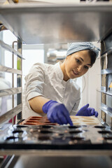 Female kitchen staff in uniform putting many rubber mold tray for cooking candy into oven or shelves