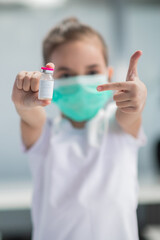 A cute masked girl holds a vaccine in her hand and points at it with her finger