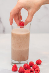 Chocolate milk shake with fresh raspberries close up on marble background, woman hands, vertical banner